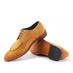 May include: Mustard yellow suede dress shoes with brown laces and a leather sole. The shoes have a classic design with a stitched welt and a rounded toe. The sole is made of dark brown leather with a black rubber heel.