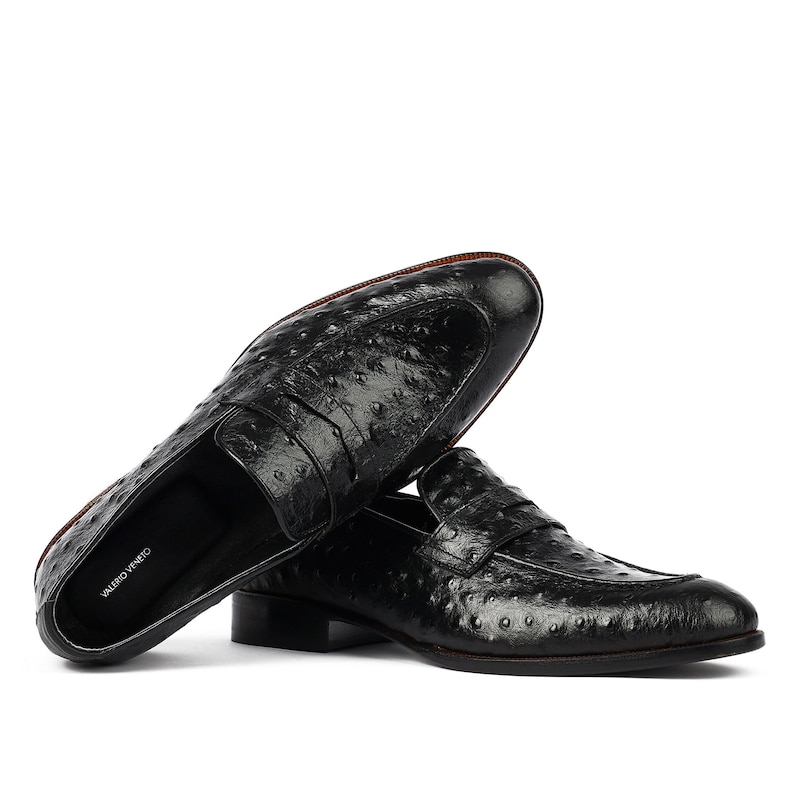 May include: Black leather loafers with an ostrich print design. The loafers have a penny strap detail and a brown leather sole. The brand name "Valerio Veneto" is visible on the insole.