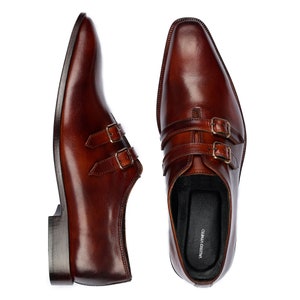 May include: A pair of brown leather double monk strap shoes with gold buckles. The shoes have a pointed toe and are made of high-quality leather.