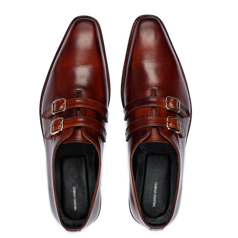 May include: A pair of brown leather double buckle monk strap shoes. The shoes have a pointed toe and are made of a smooth leather. The brand name "VALERIO VENEITO" is printed on the inside of each shoe.