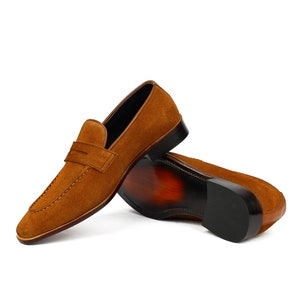 May include: A pair of brown suede loafers with a leather sole. The loafers have a simple design with a single strap across the top.