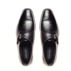 May include: A pair of black leather monk strap shoes with a silver buckle. The shoes have a pointed toe and a low heel. The brand name "VALERIO VENETO" is printed on the inside of each shoe.