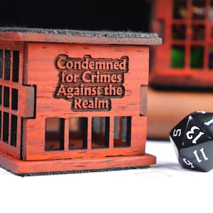 May include: A red wooden miniature jail cell with the text "Condemned for Crimes Against the Realm" on the front. A black 20-sided die with white numbers is in the foreground.
