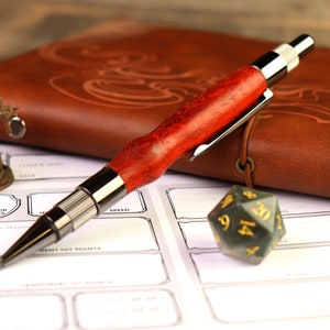 May include: A red and silver pen with a wood grain pattern on the barrel, lying on a brown leather journal with a green 20-sided die next to it.