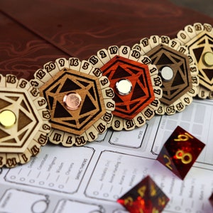 May include: Five wooden dice spinners with a metal center piece. The spinners are numbered 1-20. The dice spinners are on a white surface with a brown leather notebook in the background.