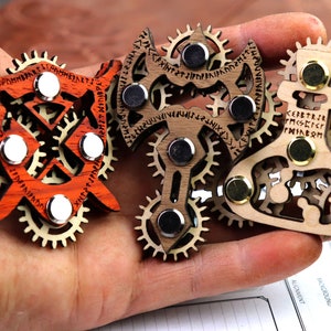 May include: Three wooden, gear-shaped, magnetic fidget toys. The first is a shield with a red and brown wood grain, the second is an axe with a brown wood grain, and the third is a flask with a brown wood grain. Each toy has runes carved into the wood.
