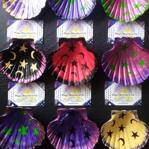 Könnte beinhalten: Neun bemalte Muscheln mit verschiedenen Stern- und Halbmond-Designs in den Farben Lila, Rosa, Gelb und Rot. Die Muscheln sind in drei Reihen zu je drei angeordnet. Jede Muschel hat ein anderes Design. Der Text "Magie Blanche & Cie" ist auf einer Karte unter jeder Reihe von Muscheln gedruckt.
