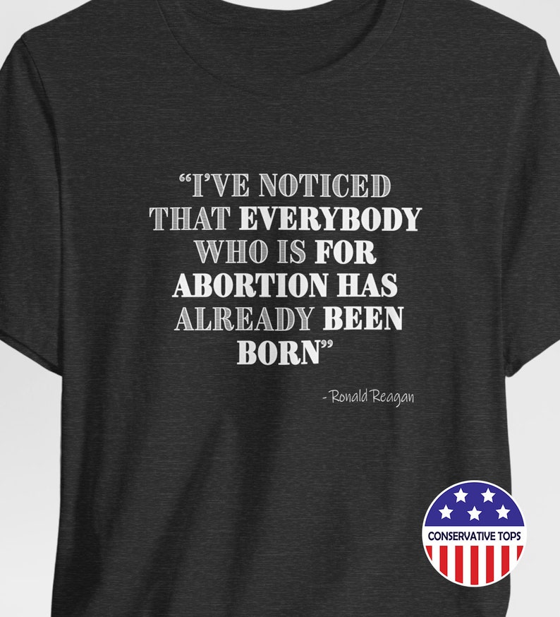 May include: A dark grey t-shirt with a white quote from Ronald Reagan that reads "I've noticed that everybody who is for abortion has already been born." The shirt also has a small American flag with the words "Conservative Tops" printed below it.