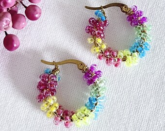 Pode incluir: Um par de brincos de argola com contas de arco-íris. Os brincos são feitos com pequenas contas coloridas que são enfiadas juntas em um padrão circular. As argolas são de metal dourado.