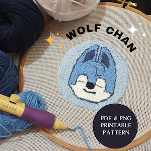 May include: A blue and white needlepoint embroidery hoop with a cartoon wolf face in the centre. The hoop is surrounded by yarn and embroidery tools. The text "WOLF CHAN" is written above the hoop. The text "PDF & PNG PRINTABLE PATTERN" is written below the hoop.