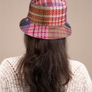 May include: A bucket hat featuring a multi-colored plaid pattern with shades of pink, orange, green, and blue. The hat has a textured, woven fabric construction with a rounded crown and a downward-sloping brim that encircles the head.