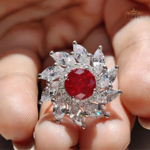 May include: A silver ring featuring a large red gemstone encircled by smaller white gemstones, arranged in a floral pattern.