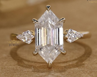 May include: A gold ring featuring a large, clear, hexagonal-cut gemstone as the centerpiece, flanked by two smaller pear-shaped stones. The ring has a delicate band and is set against a neutral background.