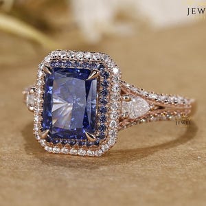 Op de afbeelding: Een roségouden ring met een grote, rechthoekige, blauwe edelsteen in het midden. De edelsteen is omgeven door een halo van kleinere witte en blauwe stenen. De ringband heeft extra kleine stenen en een decoratief ontwerp.
