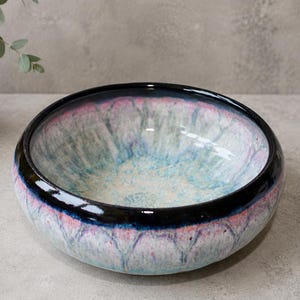 May include: A round ceramic bowl featuring a glossy black rim that transitions into a textured, mottled glaze of soft pink, light blue, and white. The interior and exterior surfaces display a reactive finish with subtle vertical drip patterns.