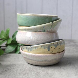 May include: A set of three ceramic mugs with a speckled, matte finish. The mugs are in shades of cream, light brown, and dark brown. Each mug has a simple, minimalist design and a rounded handle.