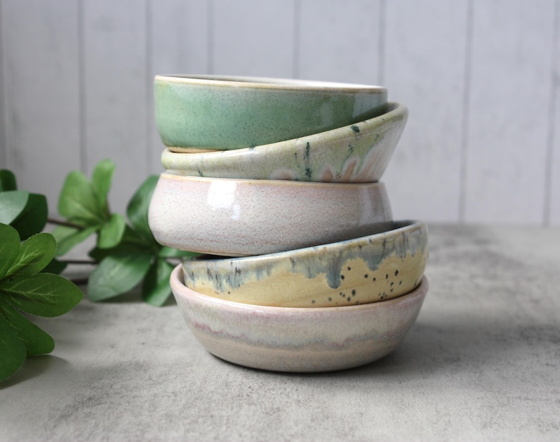 May include: A set of three ceramic mugs with a speckled, matte finish. The mugs are in shades of cream, light brown, and dark brown. Each mug has a simple, minimalist design and a rounded handle.