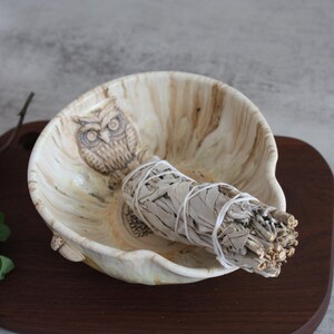 May include: A beige-toned bowl with an owl carving, containing a bundle of white sage. The bowl rests on a dark wooden surface, with green foliage in the corner. The bowl's design has a natural, earthy aesthetic, with a focus on the sage.