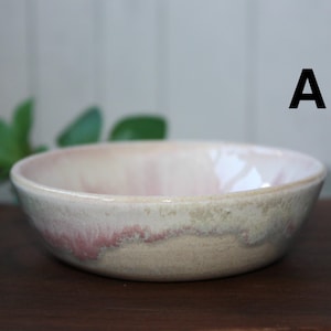 May include: A ceramic bowl with a soft pink interior and a rim that fades to a creamy white and tan. The bowl has a simple, rounded shape and is set against a dark wooden surface.