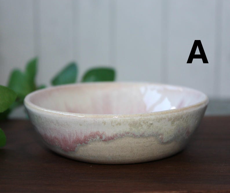 May include: A ceramic bowl with a soft pink interior and a rim that fades to a creamy white and tan. The bowl has a simple, rounded shape and is set against a dark wooden surface.