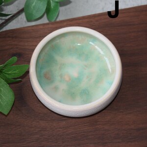 May include: A small, round ceramic dish with a white exterior and a marbled interior of green, blue, and beige hues. The dish is set on a dark wooden surface, with green leaves in the background. The letter "J" is visible.