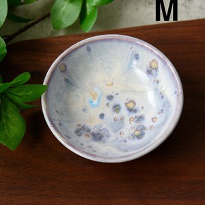 May include: A small, round ceramic bowl with a light blue and white glaze. The bowl has a speckled pattern with hints of yellow and purple. The rim is a darker shade of purple. The bowl is sitting on a dark brown wooden surface.