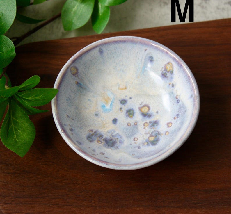 May include: A small, round ceramic bowl with a light blue and white glaze. The bowl has a speckled pattern with hints of yellow and purple. The rim is a darker shade of purple. The bowl is sitting on a dark brown wooden surface.