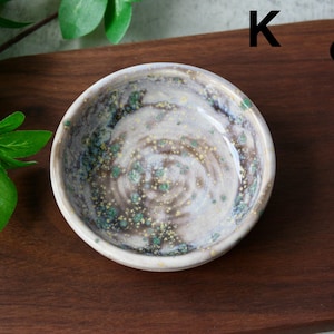 May include: A small, round ceramic bowl with a speckled, glazed interior. The bowl features a swirling pattern of brown, white, and green hues. The letter "K" is visible in the upper right corner. The bowl is set on a dark wooden surface.