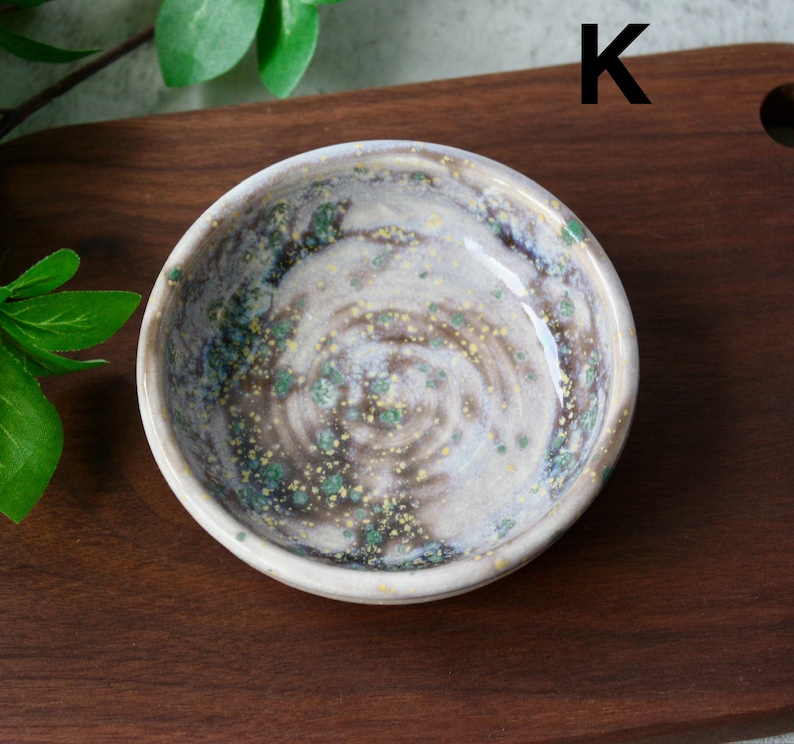 May include: A small, round ceramic bowl with a speckled, glazed interior. The bowl features a swirling pattern of brown, white, and green hues. The letter "K" is visible in the upper right corner. The bowl is set on a dark wooden surface.