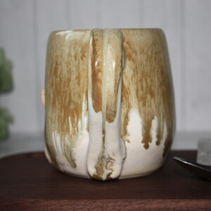 Pode incluir: Uma caneca de cer&acirc;mica artesanal com uma vidragem &uacute;nica. A caneca tem um formato arredondado com uma al&ccedil;a grande. A vidragem apresenta uma mistura de cores castanhas e creme, com um efeito de gotejamento. A caneca est&aacute; sobre uma superf&iacute;cie de madeira.