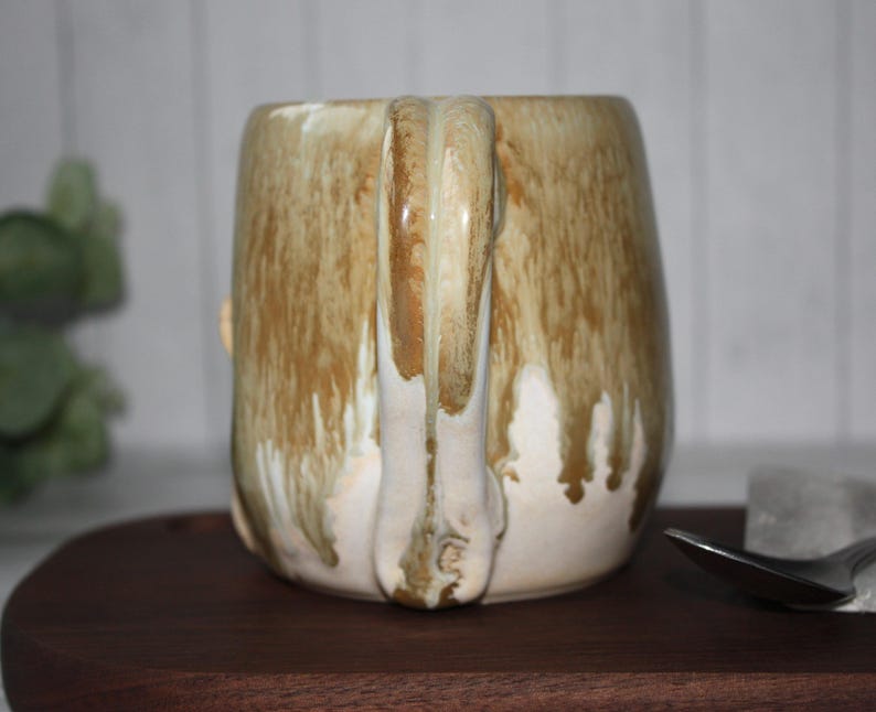 Pode incluir: Uma caneca de cer&acirc;mica artesanal com uma vidragem &uacute;nica. A caneca tem um formato arredondado com uma al&ccedil;a grande. A vidragem apresenta uma mistura de cores castanhas e creme, com um efeito de gotejamento. A caneca est&aacute; sobre uma superf&iacute;cie de madeira.