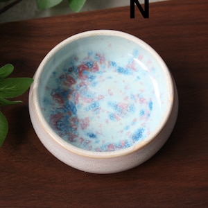 May include: A small, round ceramic bowl with a light pink exterior and a speckled interior. The interior features a light blue base with red and blue speckles. The bowl is set on a dark wooden surface.