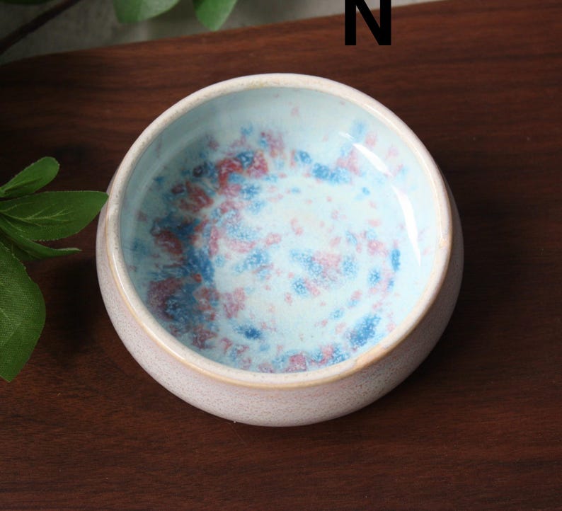 May include: A small, round ceramic bowl with a light pink exterior and a speckled interior. The interior features a light blue base with red and blue speckles. The bowl is set on a dark wooden surface.