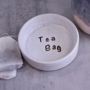 May include: A white ceramic tea bag holder with the words "Tea Bag" in black lettering. The holder has a speckled design and is next to two tea bags. A blue and white ceramic mug is in the background.