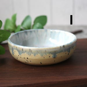 May include: A small, handmade ceramic bowl with a speckled, light yellow exterior and a blue-grey rim. The interior is a pale blue. The bowl sits on a dark wooden surface, with green leaves in the background.