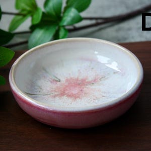 May include: A small, round ceramic bowl with a glossy finish. The bowl features a white interior with a pink floral design and a red exterior. The rim is a light beige color. The bowl is likely used for serving small portions of food or as a decorative item.