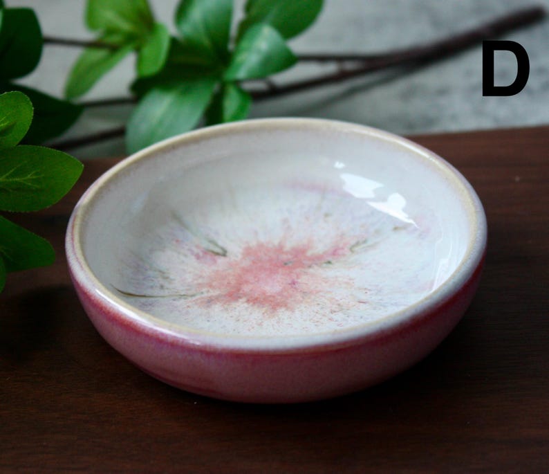May include: A small, round ceramic bowl with a glossy finish. The bowl features a white interior with a pink floral design and a red exterior. The rim is a light beige color. The bowl is likely used for serving small portions of food or as a decorative item.
