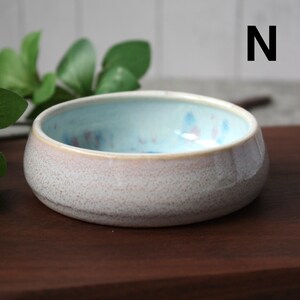 May include: A small, round ceramic bowl with a speckled, off-white exterior and a light blue interior. The rim has a soft pink hue. The bowl is sitting on a dark brown wooden surface. The letter "N" is visible in the background.