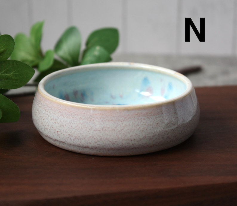 May include: A small, round ceramic bowl with a speckled, off-white exterior and a light blue interior. The rim has a soft pink hue. The bowl is sitting on a dark brown wooden surface. The letter "N" is visible in the background.