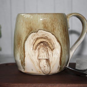 Pode incluir: Uma caneca de cer&acirc;mica bege com um design texturizado &uacute;nico. A caneca apresenta uma forma org&acirc;nica esculpida na frente, com uma al&ccedil;a na lateral. Est&aacute; colocada sobre uma superf&iacute;cie de madeira, com uma colher e um cubo de gelo por perto.