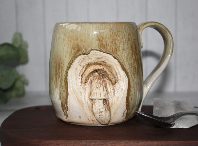 Pode incluir: Uma caneca de cer&acirc;mica bege com um design texturizado &uacute;nico. A caneca apresenta uma forma org&acirc;nica esculpida na frente, com uma al&ccedil;a na lateral. Est&aacute; colocada sobre uma superf&iacute;cie de madeira, com uma colher e um cubo de gelo por perto.