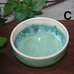 May include: A set of three ceramic mugs with a speckled, cream-colored glaze. Each mug features a different hand-painted design: a blue and green leaf, a yellow flower, and a red berry. The mugs have a simple, modern design.