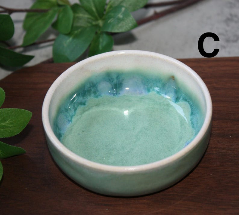 May include: A set of three ceramic mugs with a speckled, cream-colored glaze. Each mug features a different hand-painted design: a blue and green leaf, a yellow flower, and a red berry. The mugs have a simple, modern design.