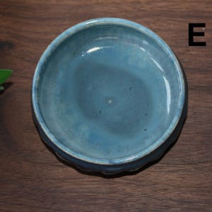 May include: A small, round, ceramic bowl with a light blue glaze. The bowl has a scalloped edge and a smooth interior. The letter "E" is visible in the upper right corner. The bowl is on a dark wood surface.