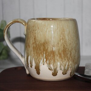 Pode incluir: Uma caneca de cer&acirc;mica artesanal com uma vidragem &uacute;nica. A caneca apresenta um corpo arredondado e uma al&ccedil;a grande. A vidragem transita de um bege claro na base para um castanho terroso rico no topo, com um efeito de gotejamento.