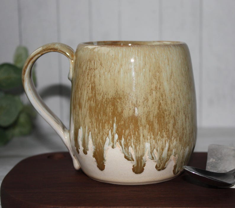 Pode incluir: Uma caneca de cer&acirc;mica artesanal com uma vidragem &uacute;nica. A caneca apresenta um corpo arredondado e uma al&ccedil;a grande. A vidragem transita de um bege claro na base para um castanho terroso rico no topo, com um efeito de gotejamento.
