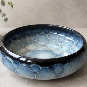 May include: A round ceramic bowl featuring a deep blue and light blue glaze with a distinctive bubble-like pattern. The rim is finished in a glossy black glaze, and the interior displays a mottled, textured appearance with shades of white and blue.