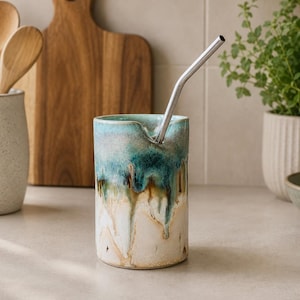 May include: A cylindrical ceramic tumbler featuring a reactive glaze with a gradient of teal, blue, and brown drips over a cream-coloured base. The vessel has a notched rim and contains a single, angled, reusable stainless steel drinking straw.
