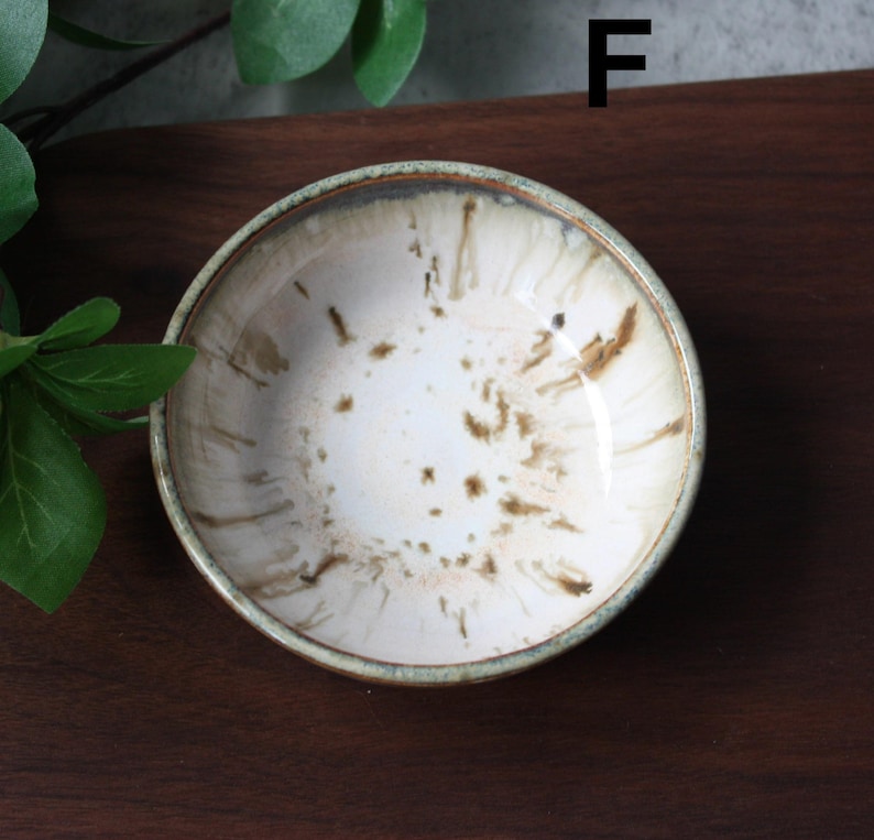 May include: A small, round ceramic bowl with a white interior and brown speckled design. The rim has a light green and brown border. The bowl is likely used for serving small portions of food or as a decorative piece.