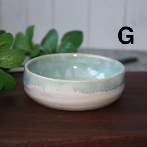 May include: A small, handmade ceramic bowl with a light blue and white glaze. The bowl has a rounded shape and a smooth, glossy finish. The letter "G" is visible in the background. The bowl is likely used for serving food or as a decorative item.